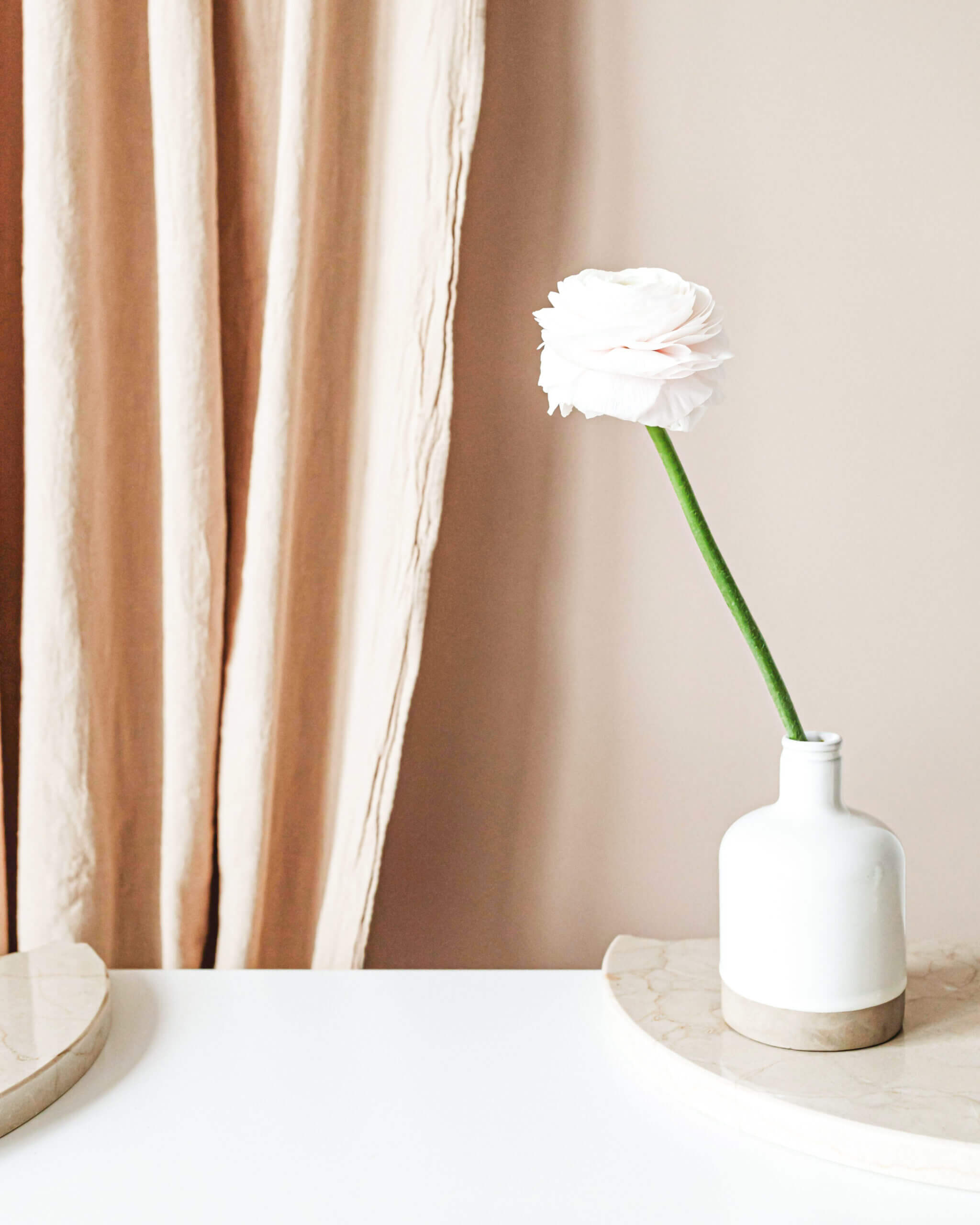 single peony stem in a white vase on a table set against a beige curtain