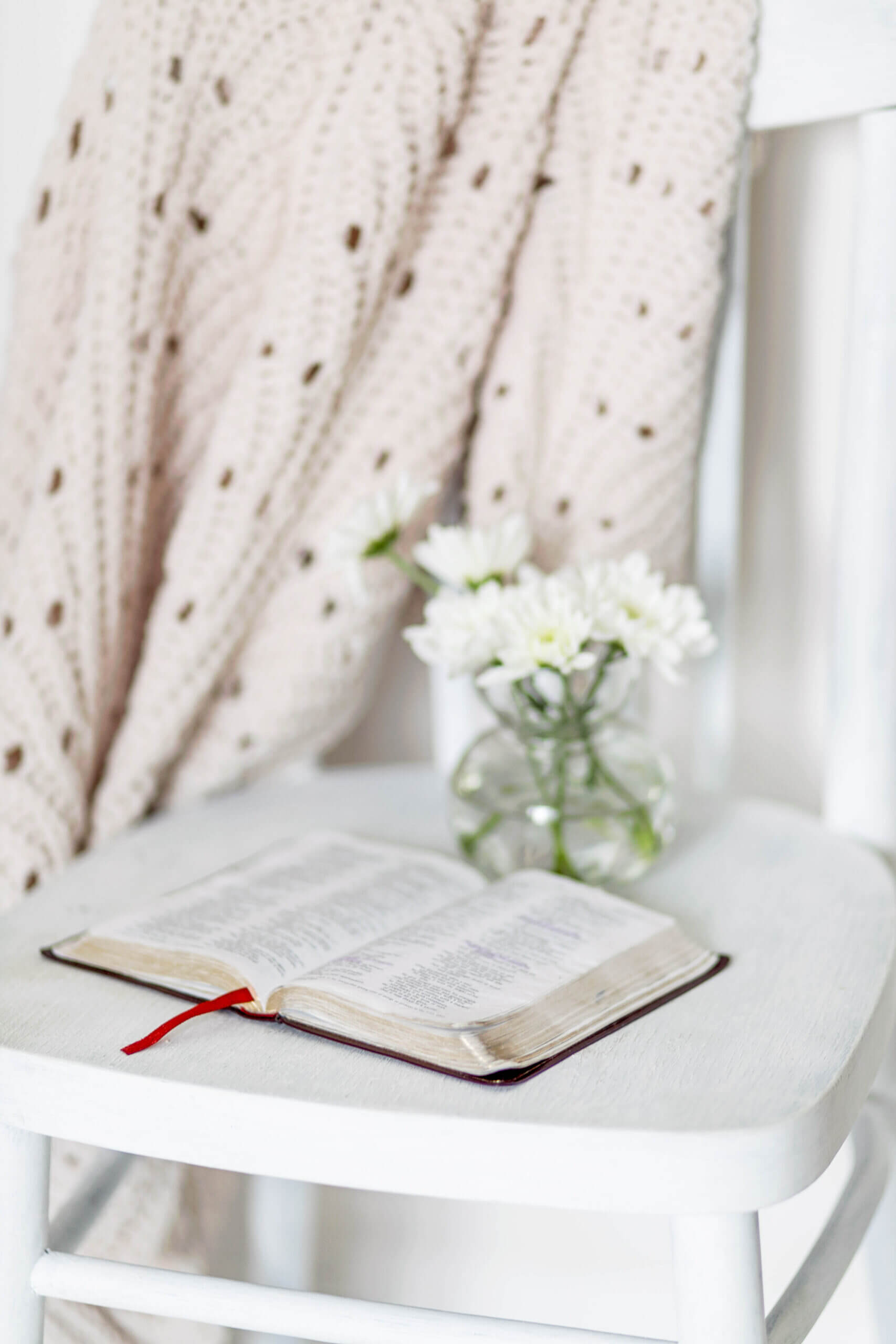 A white wooden chair with an open Bible resting on the seat, its red ribbon bookmark visible. Beside the Bible, a small glass vase holds fresh white flowers, and a cozy cream-colored knit blanket is draped over the chair's backrest, creating a peaceful and inviting atmosphere.