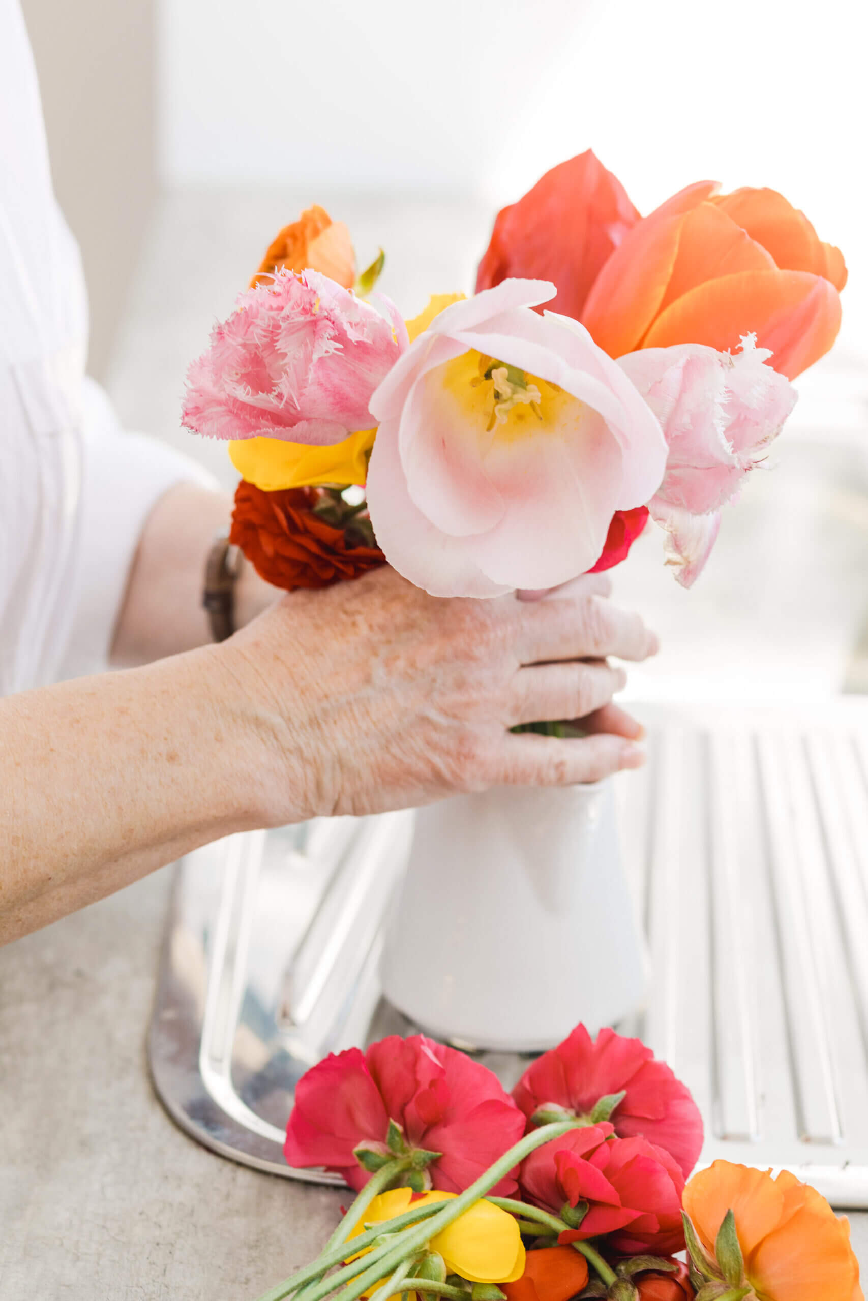 A close-up of hands arranging a bouquet of fresh tulips and ranunculus in a white vase, with additional flowers resting on a countertop